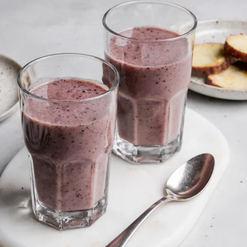 Taro root smoothie in two glasses on a kitchen table with a plate of sliced taro