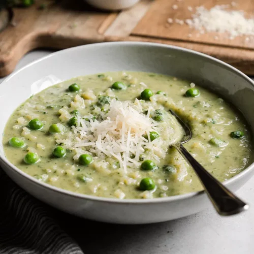 A bowl of green pea risotto served on a simple kitchen table with a fork beside it.