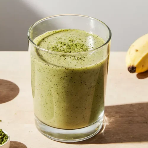 Creamy matcha protein smoothie in a glass with banana and matcha powder on a kitchen counter